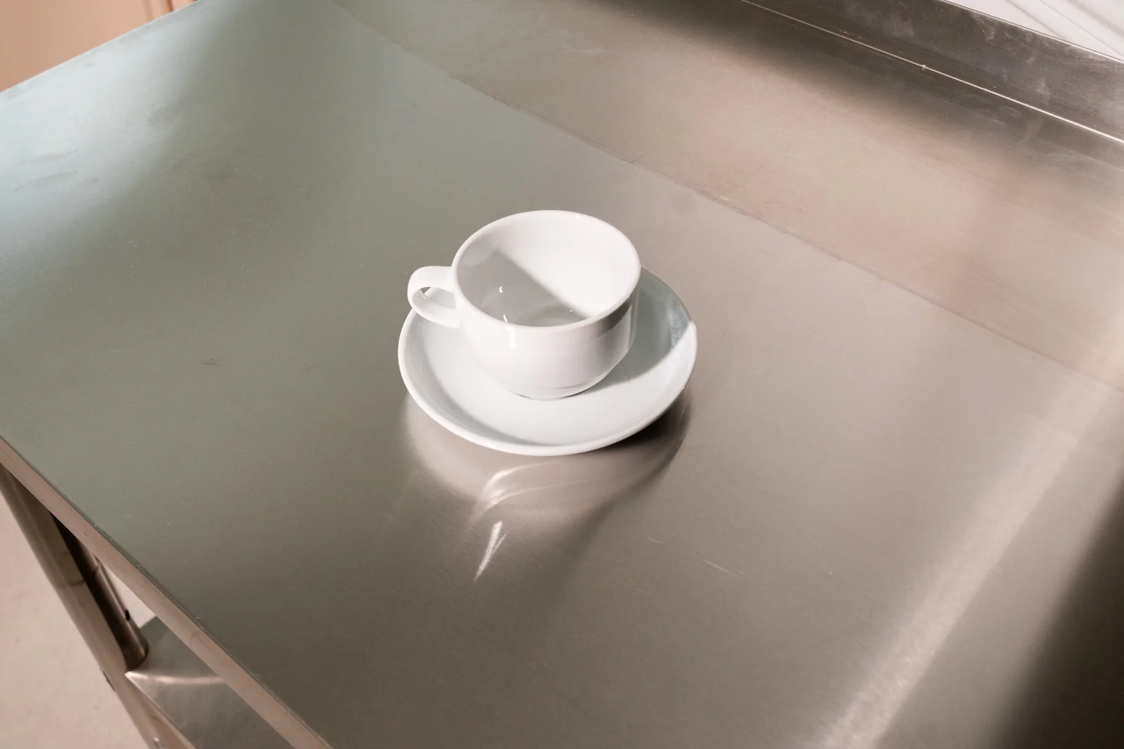 Kleine weiße Delta-Kaffeetasse mit passender Untertasse auf einer Edelstahlfläche. Weitere Ansicht.