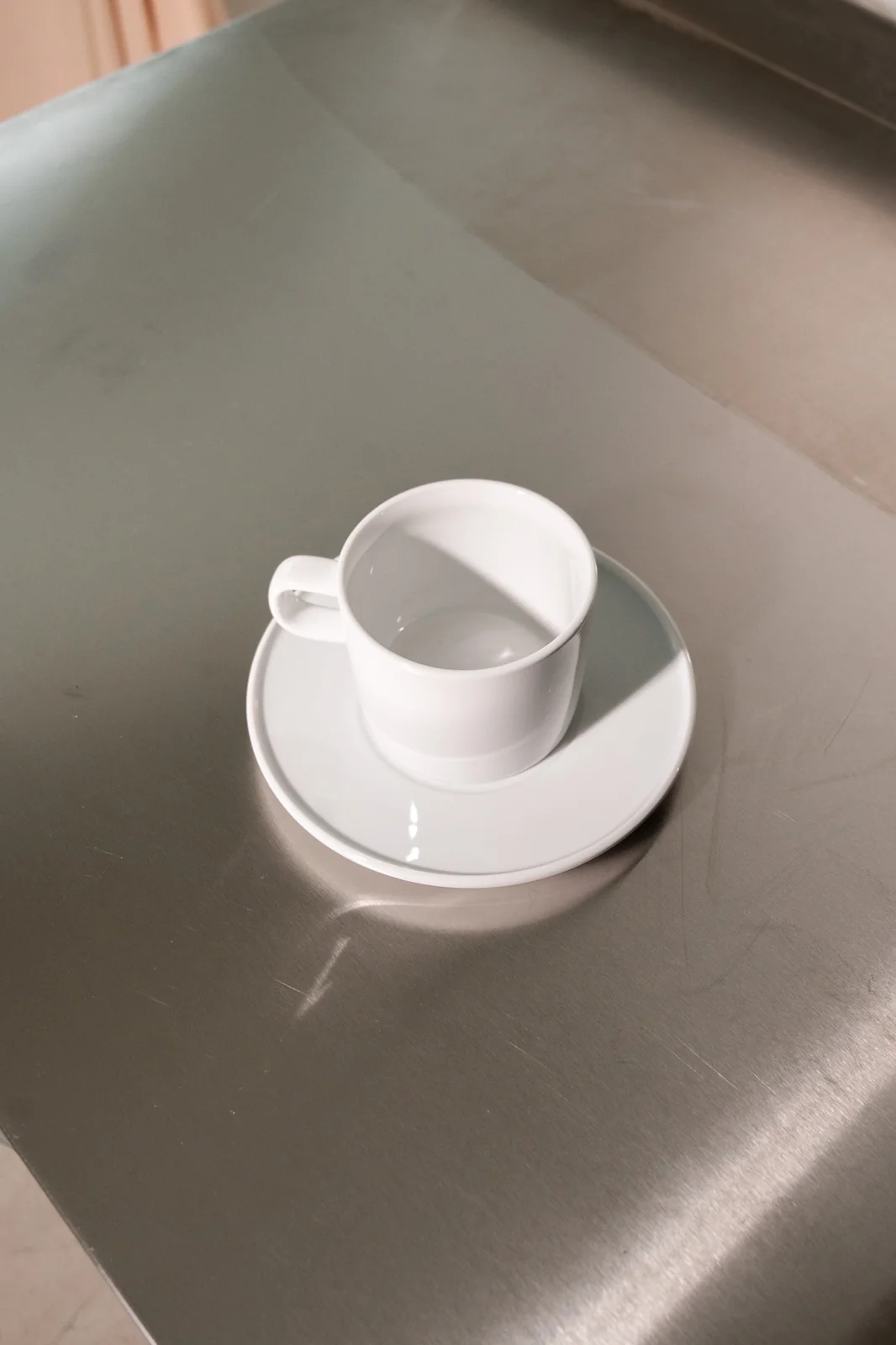 Kleine weiße Delta-Kaffeetasse mit passender Untertasse auf einer Edelstahlfläche.