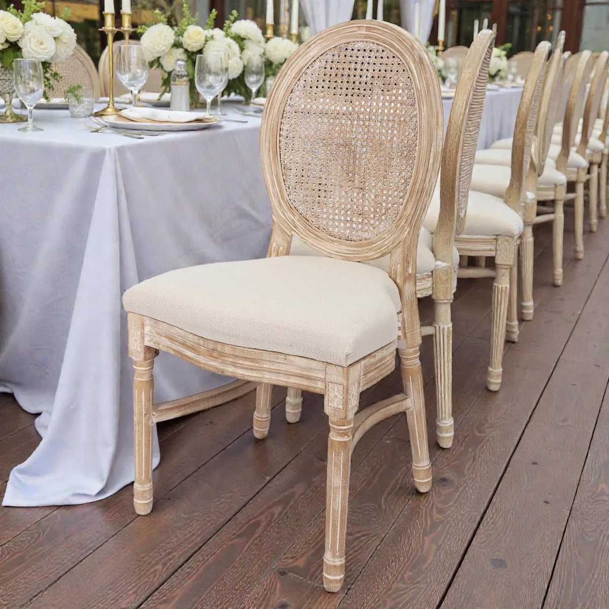Reihe von Louis-Stühlen mit ovaler Rattan-Rückenlehne und cremefarbenem Polster an einer festlich gedeckten Tafel mit weißen Rosen
